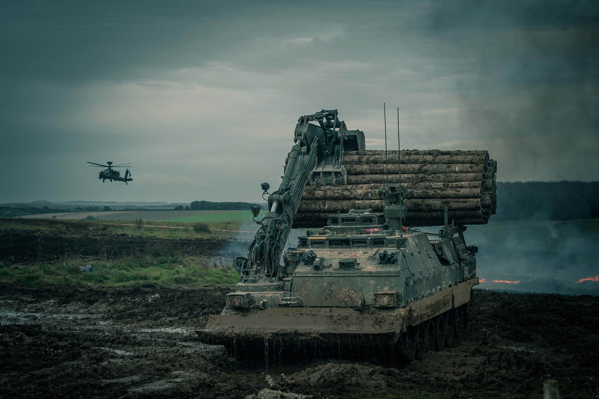 A large tank-like vehicle with a crane on the top, holding logs drives through a large muddy pit