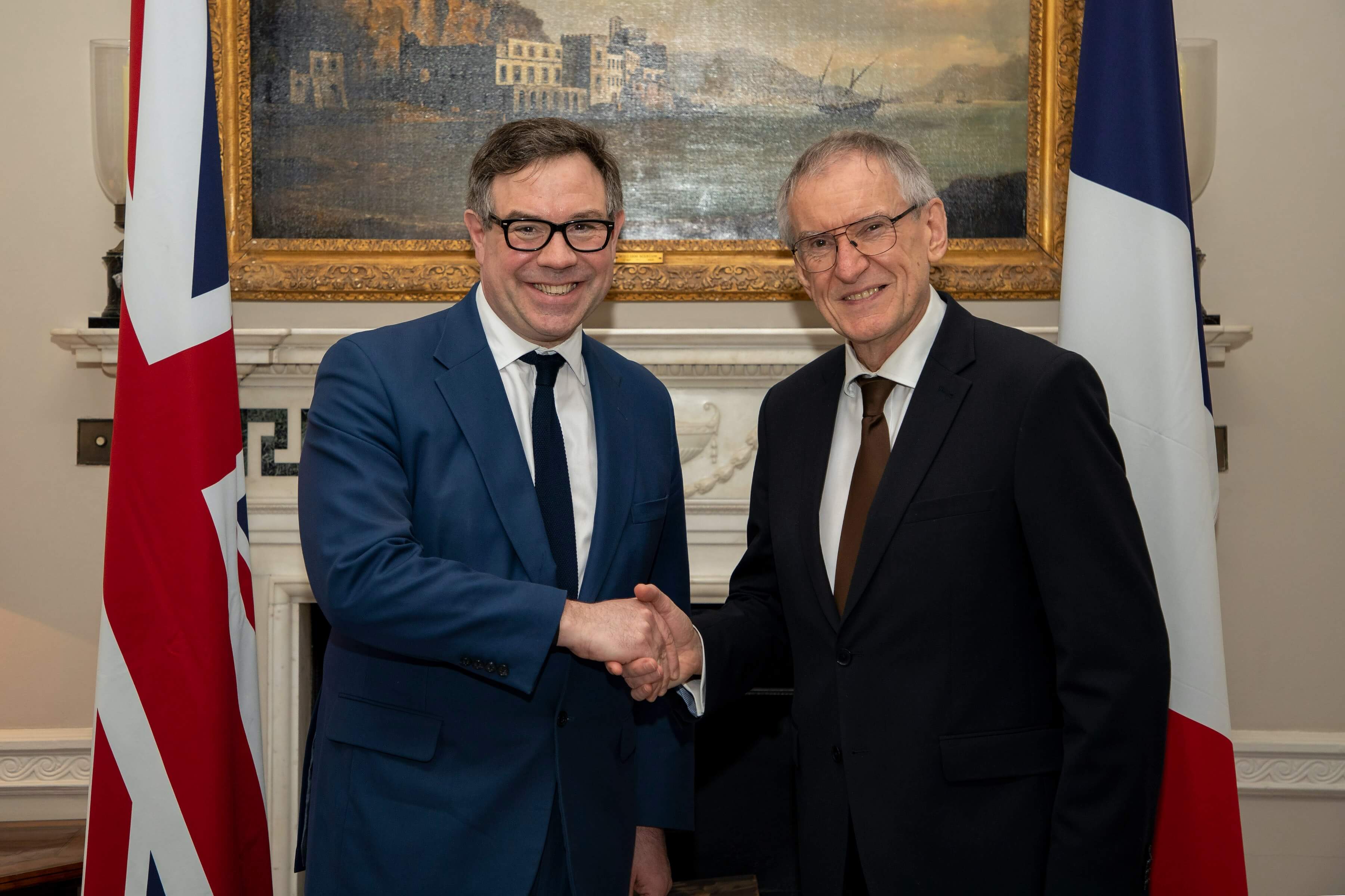 Two men wearing suits and glasses shaking hands in front of large old painting and two flags