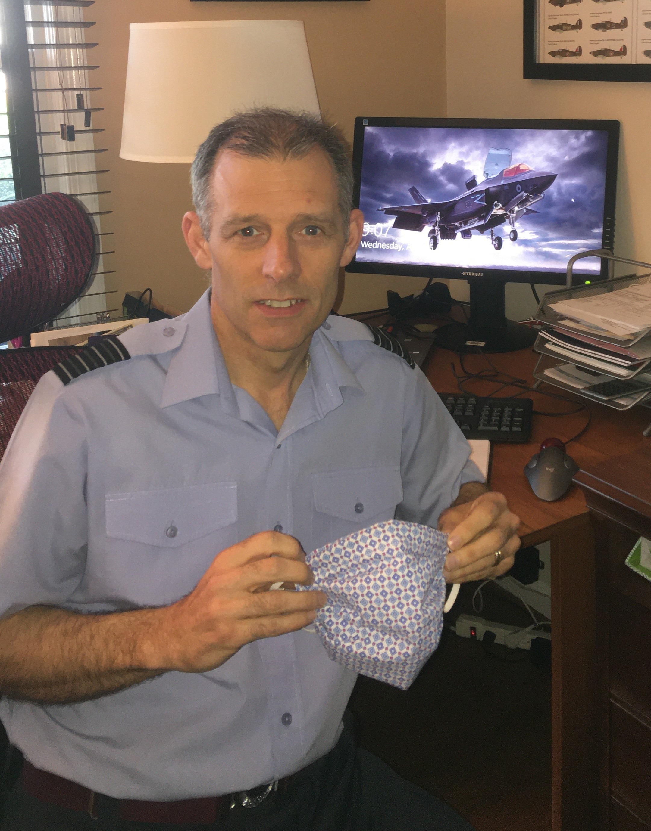 A picture of a man sat in his office wearing a light blue uniform, looking towards the camera and holding a face mask that he has made