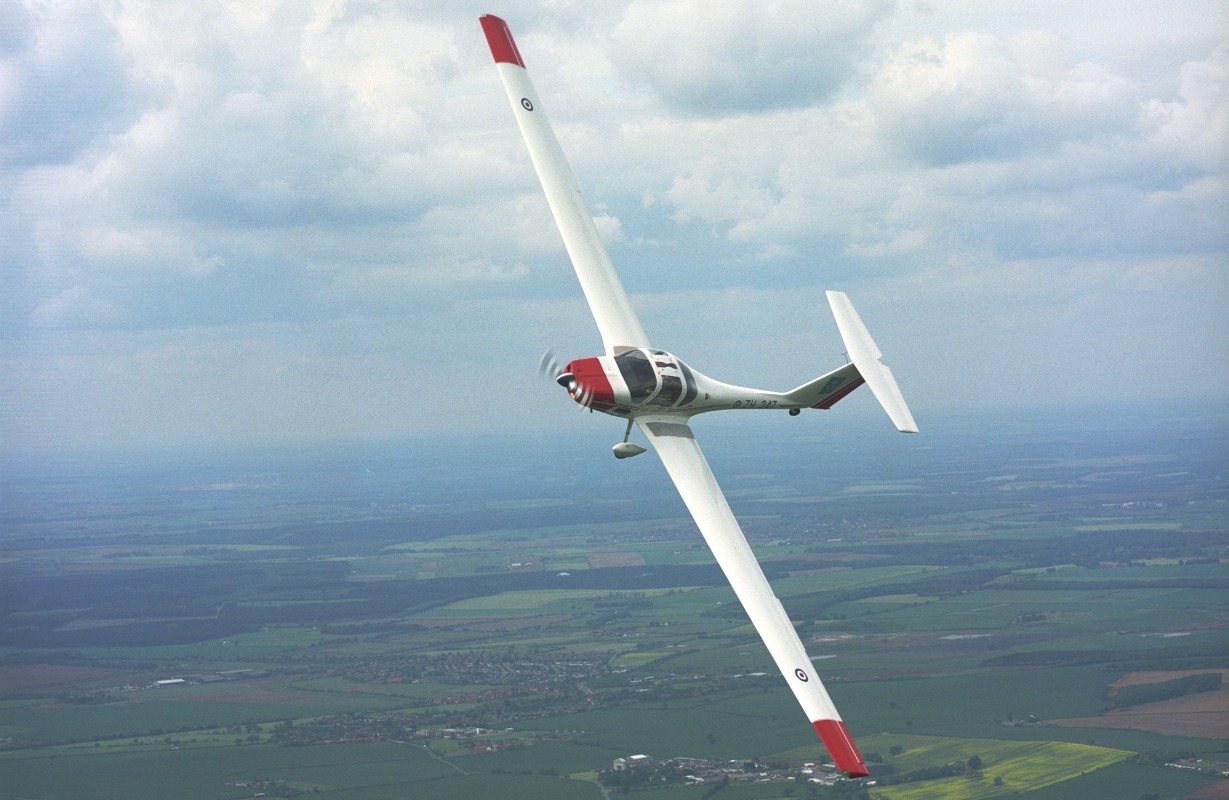 Red and white glider plane flying over green fields, dipping its wing to turn towards the camera