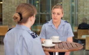 Two female royal air force personnel sat at a table with two white cups of coffee indoors at a table