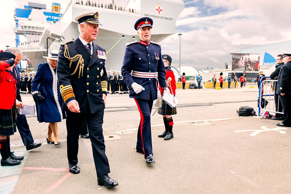 Prince Charles in navy uniform with large grey aircraft carrier in the background