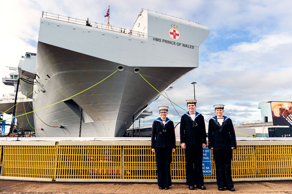 Three sailors pose in uniform in front of a large grey aircraft carrier