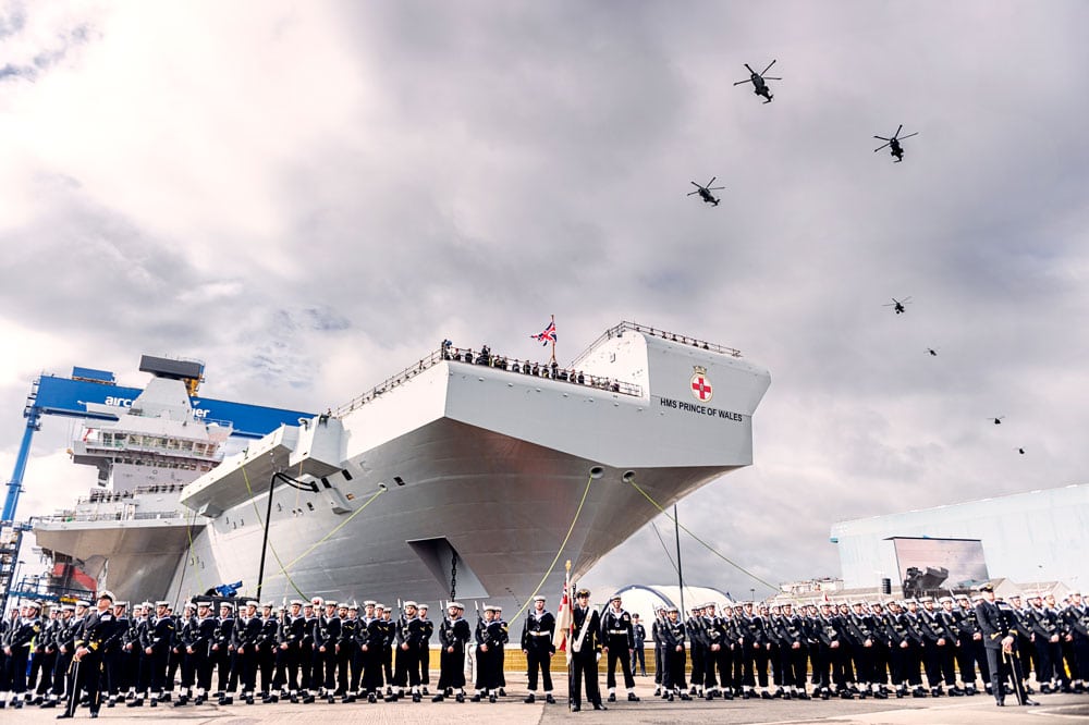 A navy marching band playing their instruments with a large aircraft carrier in the background