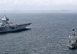 A large aircraft carrier sailing alongside a line of navy boats