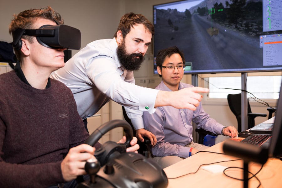 Bearded man guides someone driving in a simulator using a VR headset