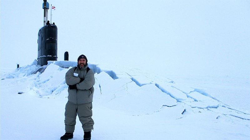 A man in winter clothing stood in front of a submarine that has broken through the arctic ice