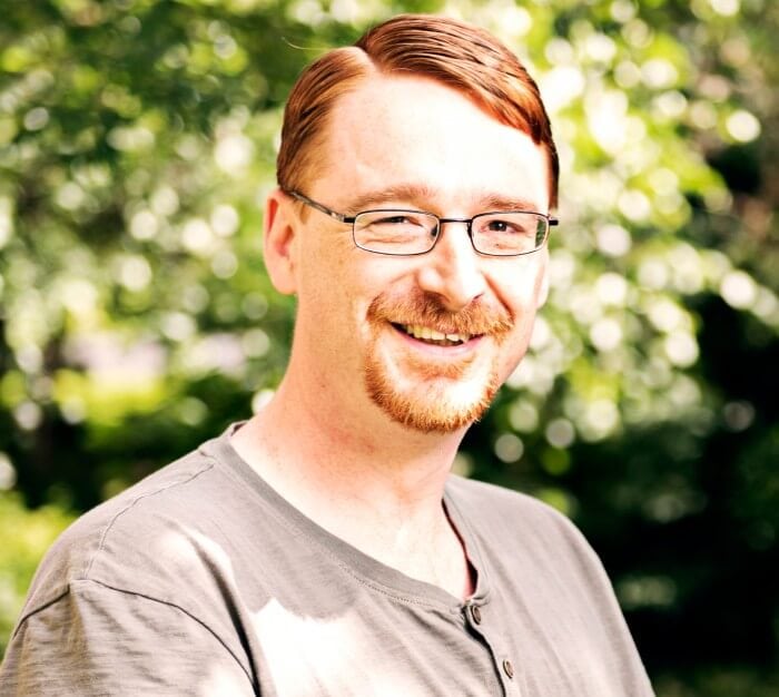 Man with ginger hair in grey t-shirt and glasses smiles at the camera