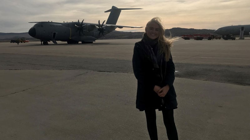 A young woman in a dark coat clasps her hands and smiles at the camera with a large grey transport plane behind her