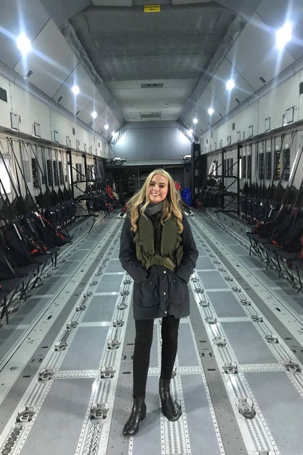 A young blonde woman smiles at the camera, her hands in the pockets of her jacket as she stands inside a large transport plane