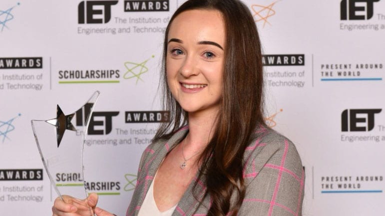 A young woman in a suit smiles at the camera against a branded background at an awards event