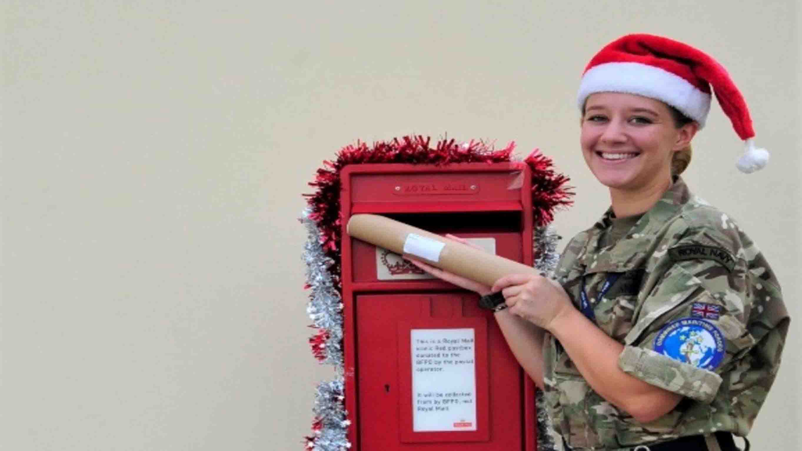 A red Royal Mail post box install in a wall in Bahrain, with a woman dressed in a Santa hair smiling as she posts a Christmas parcel