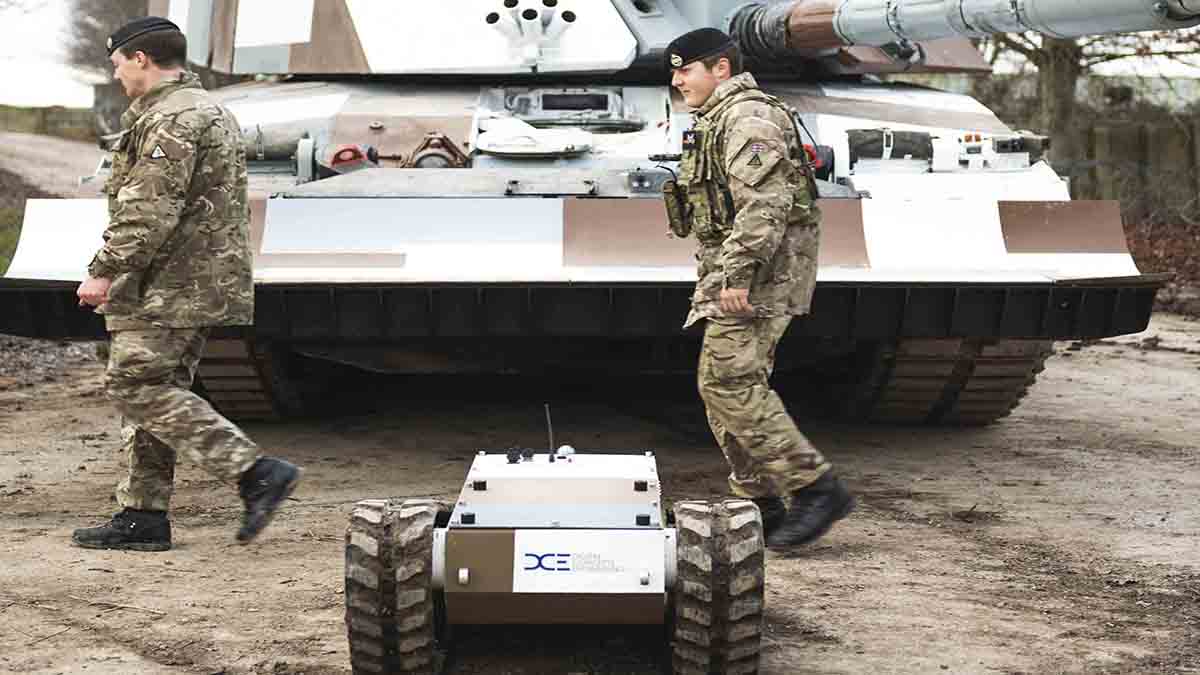 A solider stands with a small armored robot in front of a tank