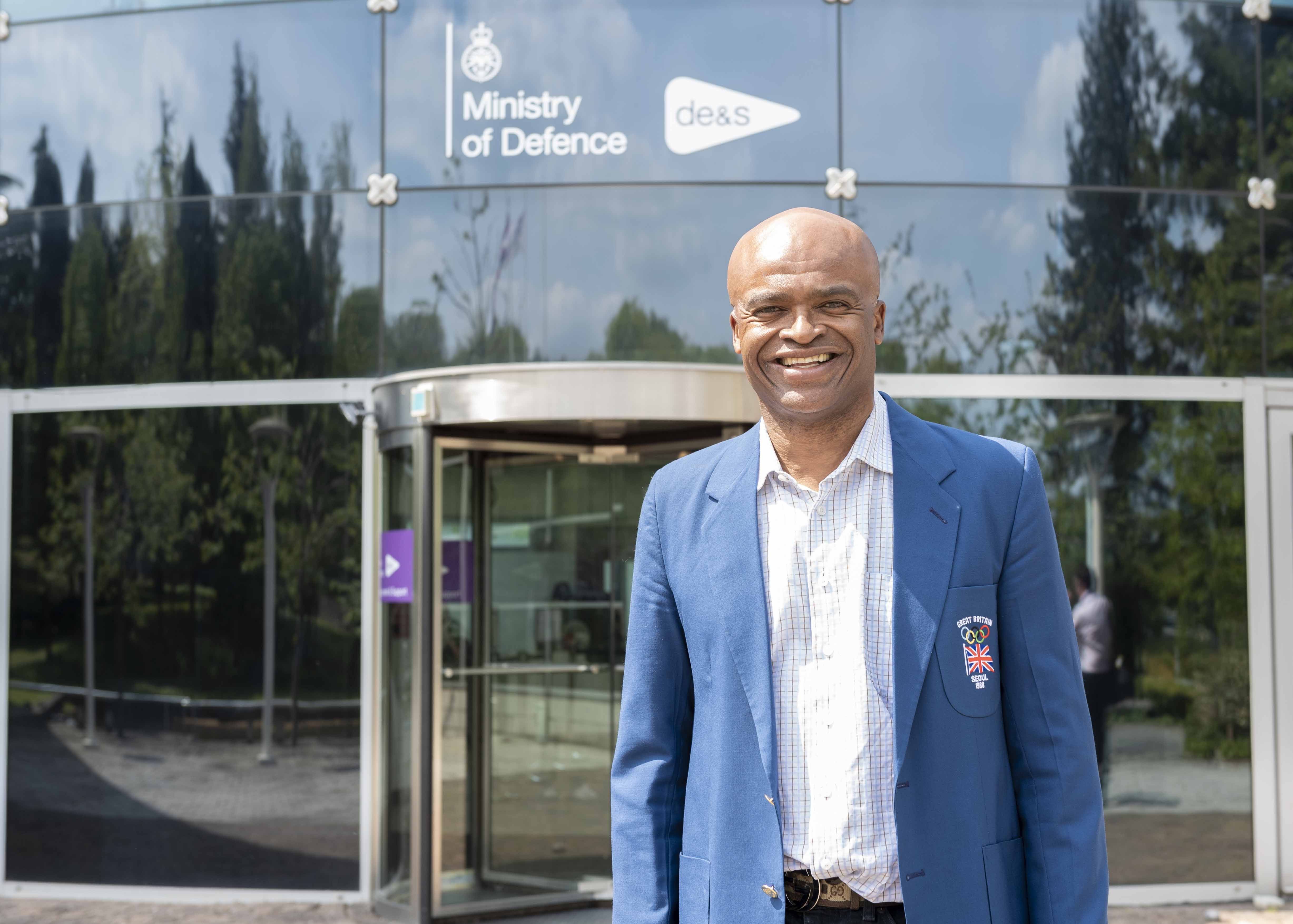 Famous athlete Kris Akabusi smiles at the camera in a blue suit as he stands outside the Defence Equipment and Support offices