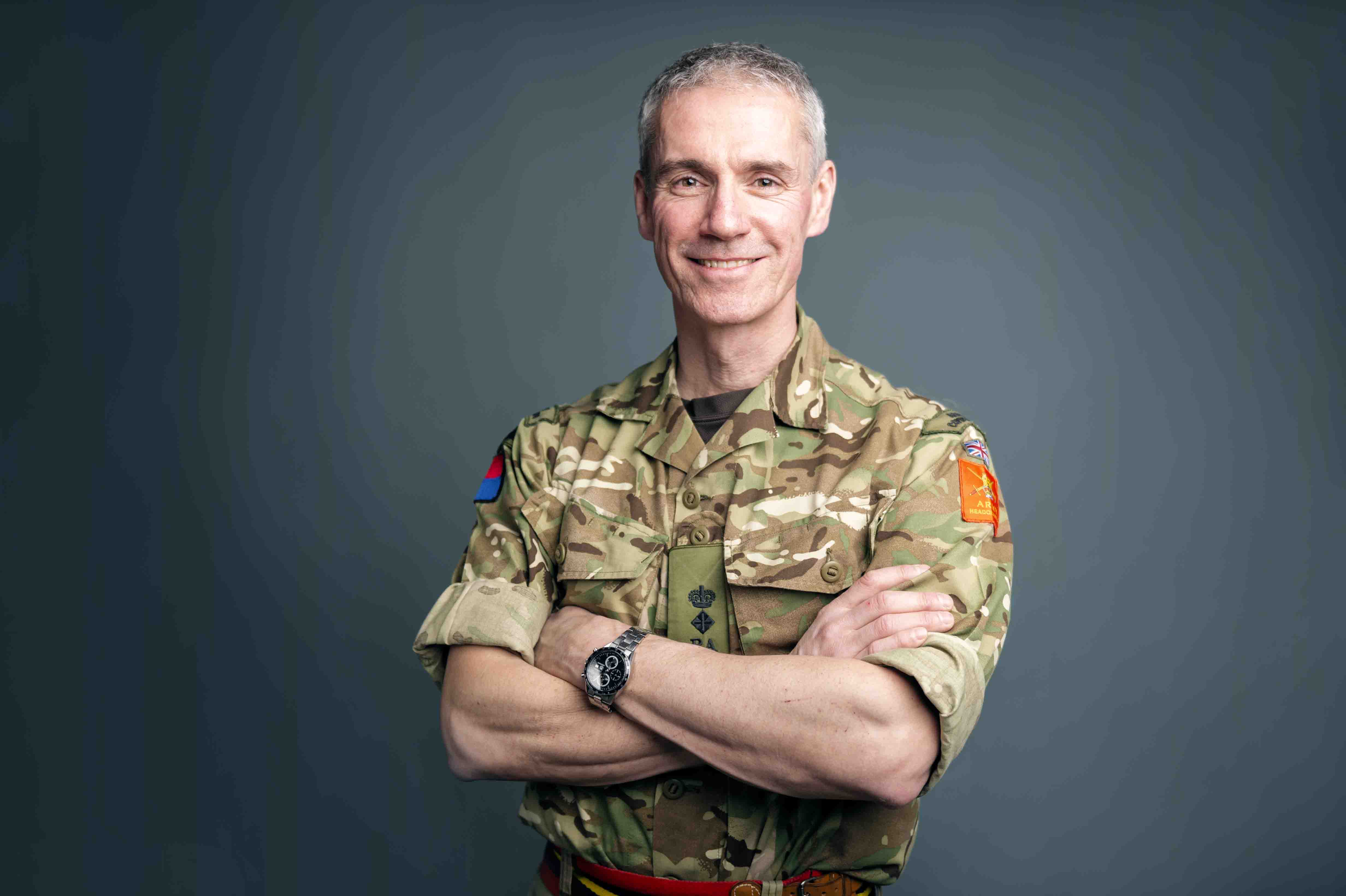 A male soldier with grey hair smiles at the camera in uniform with his arms folded