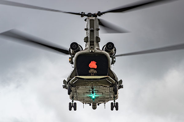 A twin rotor helicopter flying against a grey sky, with its rear door open and a solider visible inside