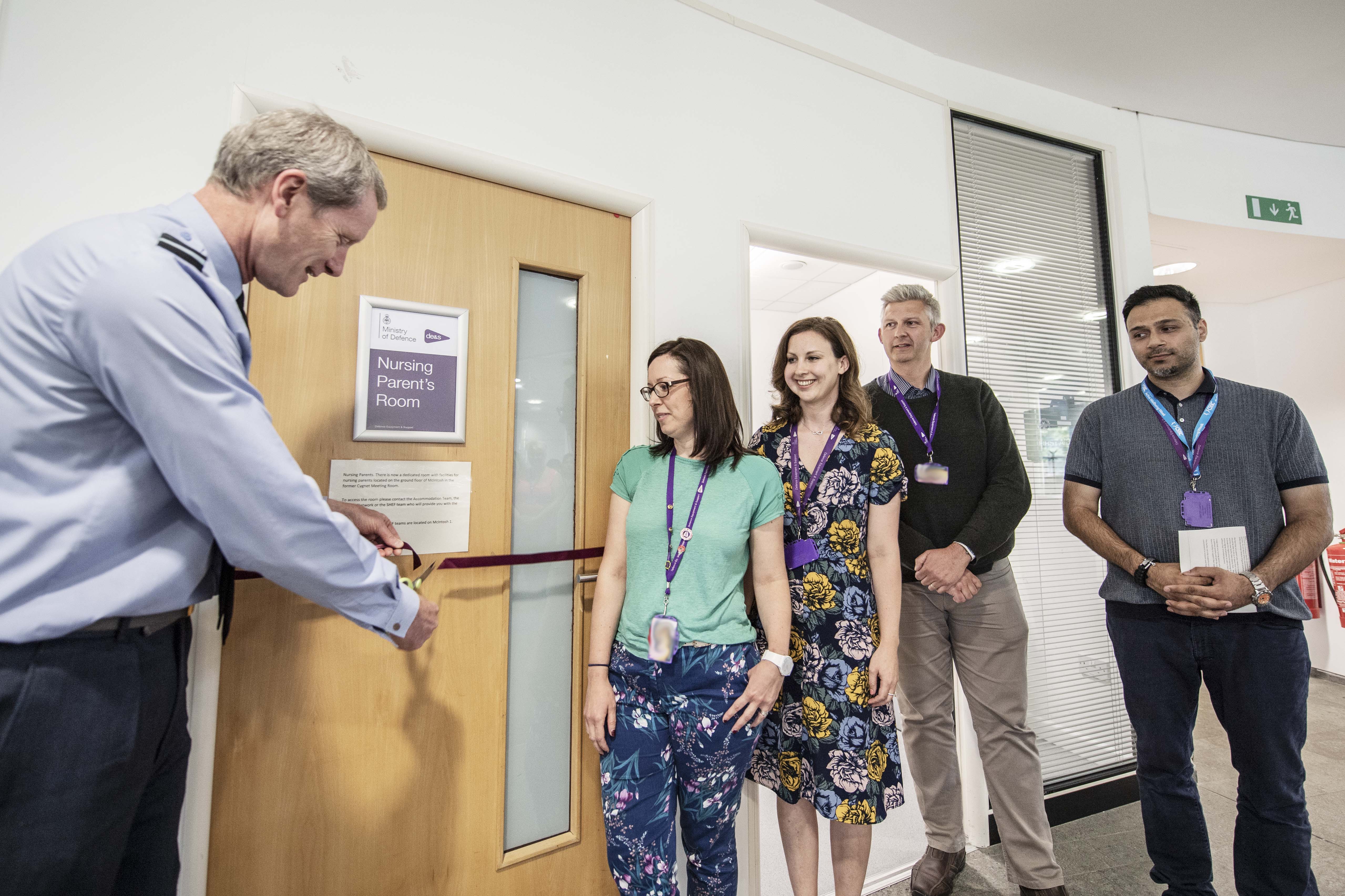 A man cuts a ribbon to open a new room in an office while a group watches on