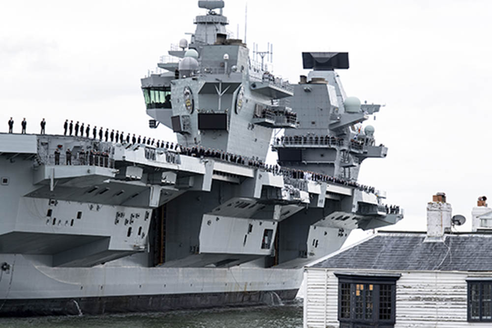 A cropped black and white photograph of a large aircraft carrier, showing its side and control tower