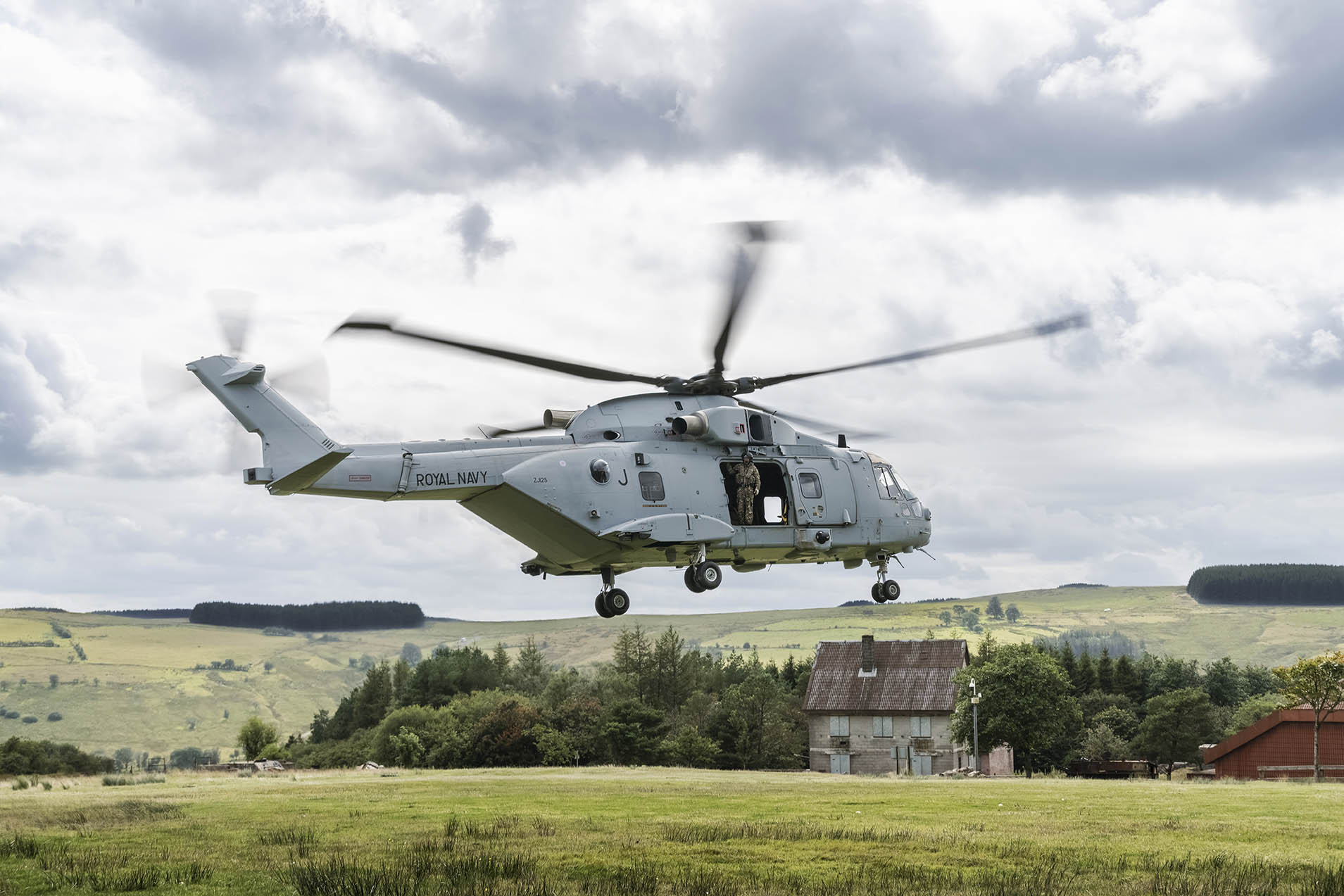 A grey helicopter about to land in a field, blowing the grass downwards