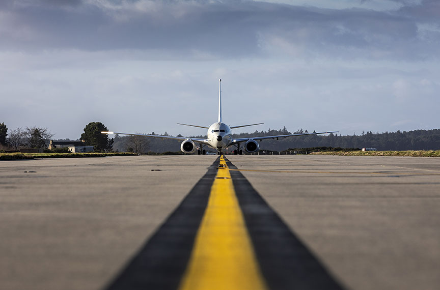 A large white plane stands on a runway, with a yellow stripe running from its front wheel to the foreground on the tarmac
