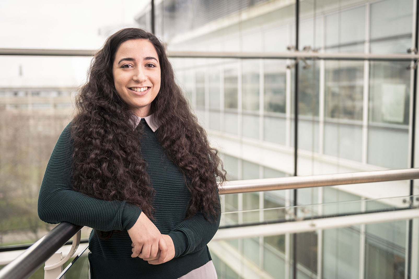 A young woman with dark hair leans on a railing inside a glass office, smiling at the camera