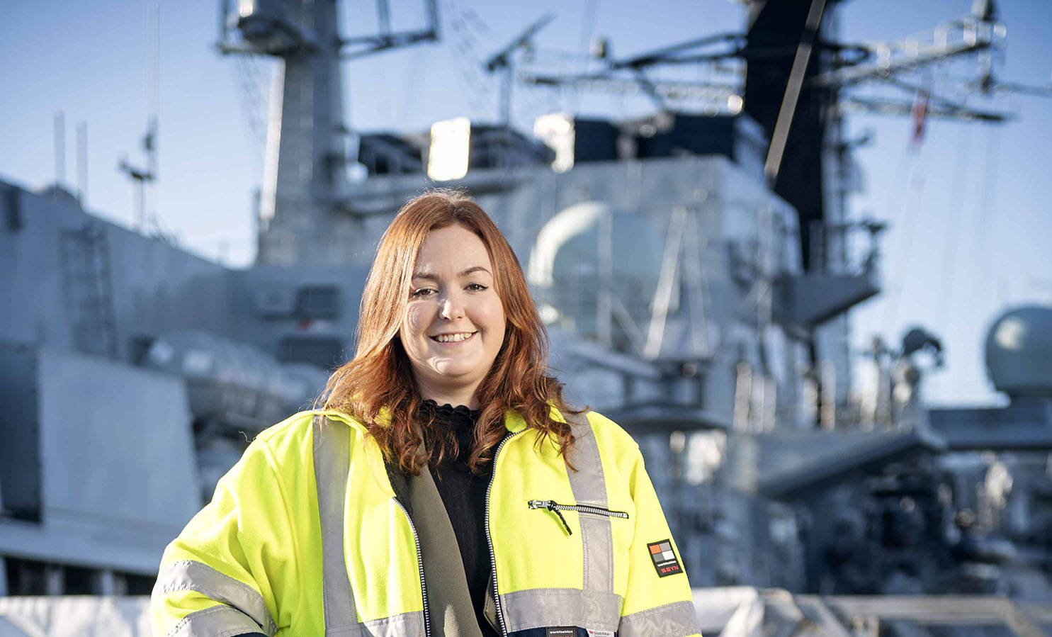 A woman in a high visibility jacket stands in front of a large grey navy ship