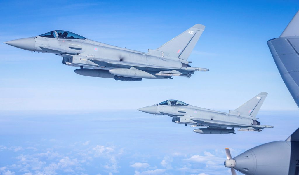 Two grey jet aircraft flying in blue skies one behind the other with the engine of another jet aircraft in the foreground