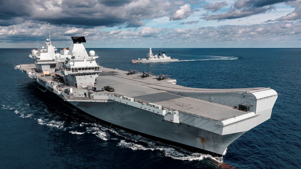 Large grey ship in the foreground with three jet aircraft on the ships deck. In the background smaller grey ship turning in the blue sea. White fluffy clouds above