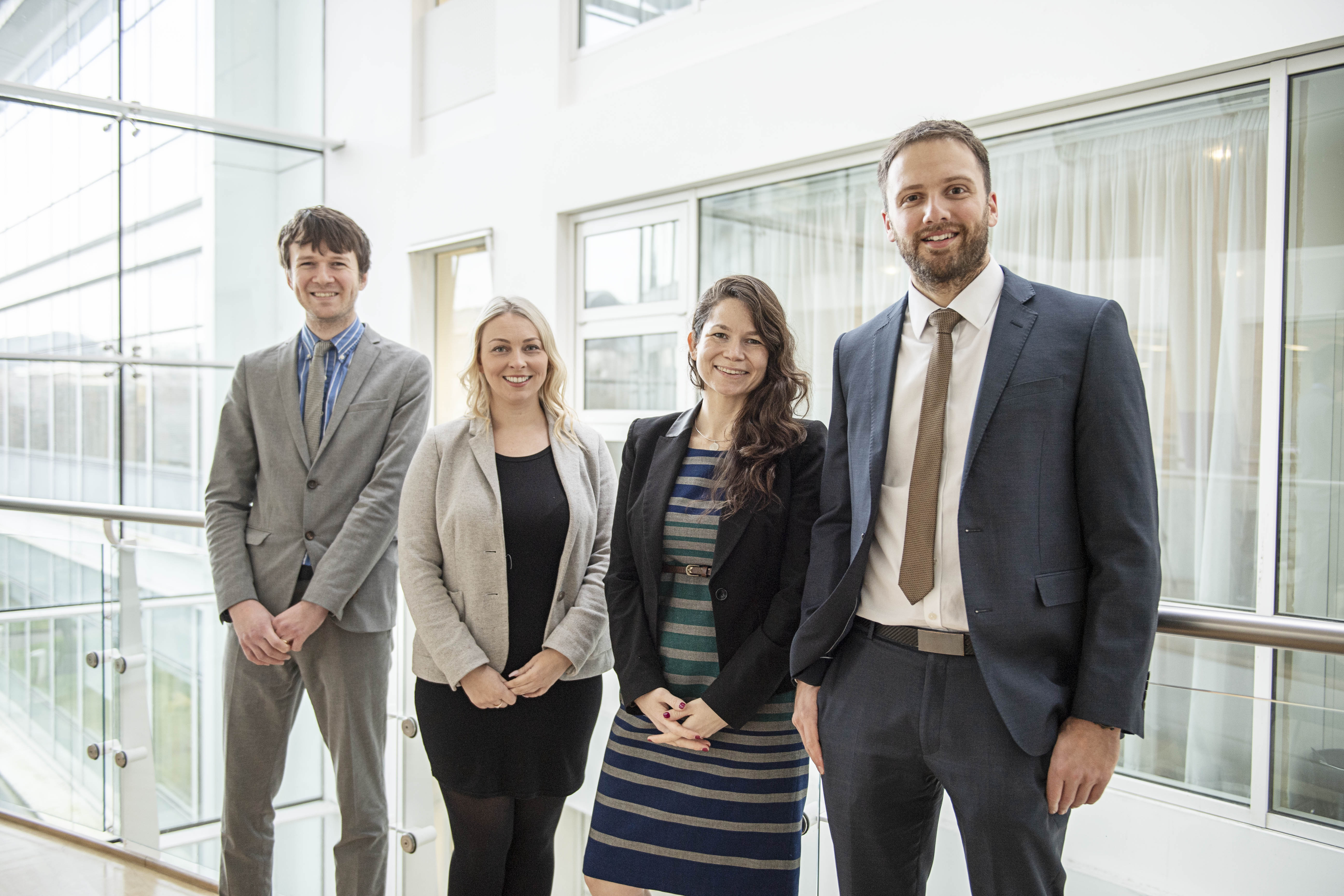 Four people in a group photo, a male on the left wearing a grey suit next to him a lady wearing business dress then another women wearing a stripy dress and black jacket. On the right a man wearing a dark blue suit. The photo has been taken indoors with windows as a background