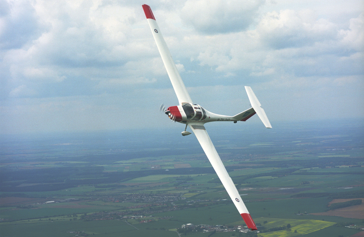 Red and white glider plane flying over green fields, dipping its wing to turn towards the camera