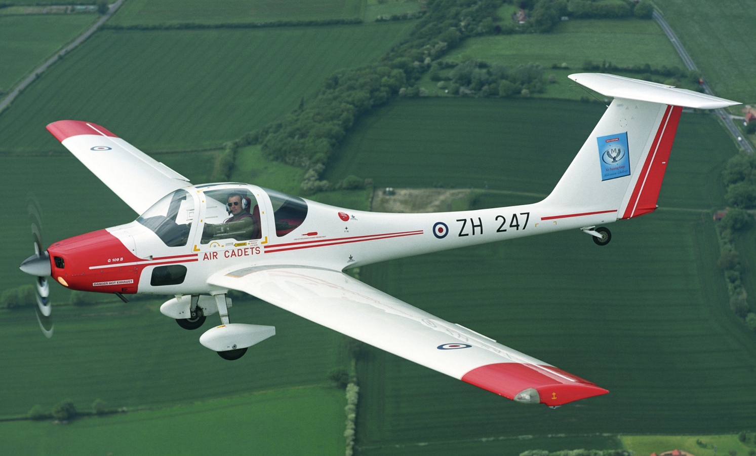 Red and white glider plane with pilot wearing green overalls flying over green fields