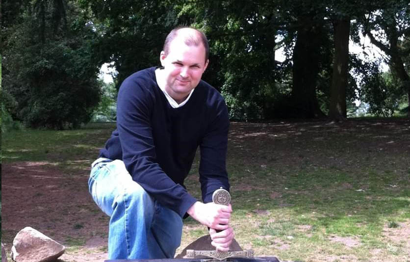 A man wearing a blue jumper and jeans kneeling down and holding the handle of a sword, the background shows green trees