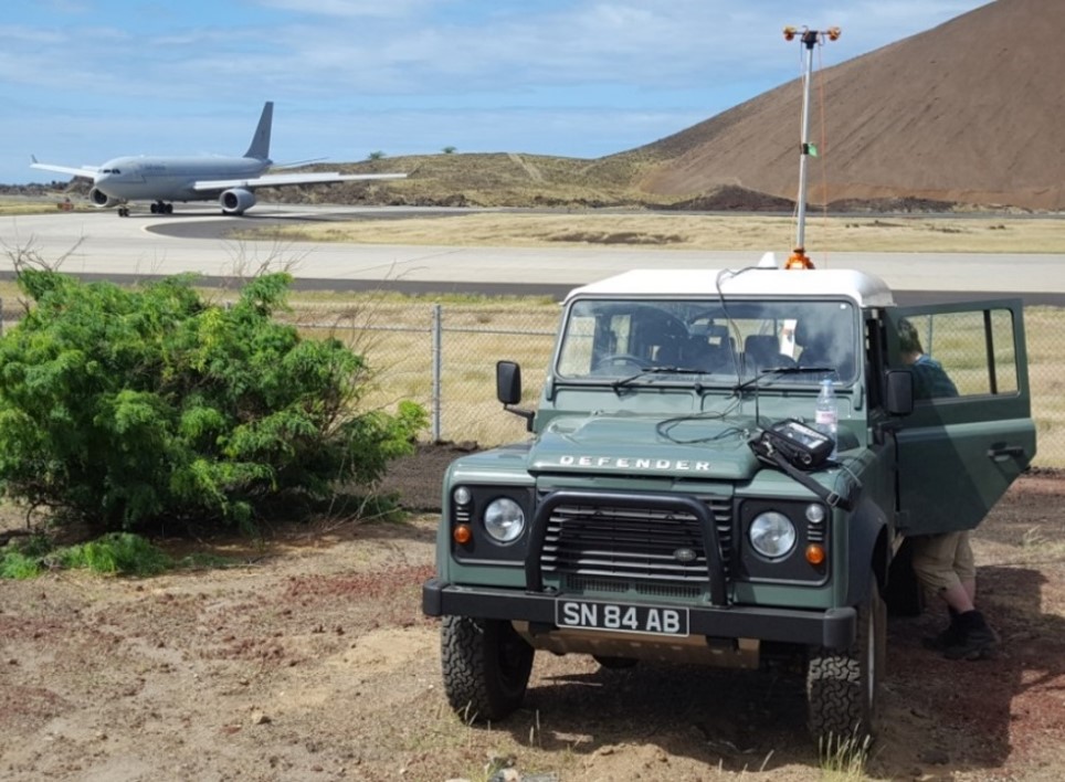 Green offroad vehicle in the foreground with an aerial on the roof. In the background a grey military aircraft parked on an airport runway in the sunshine