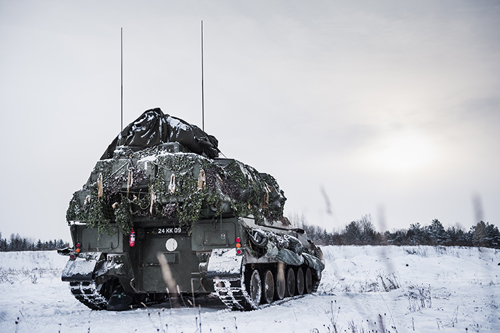 A large artillery vehicle draped in camouflage and snow drives away from the camera, through snowy terrain