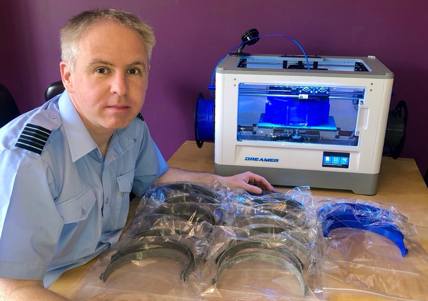 A man in light blue uniform sat in front of a 3D-printer, with various parts on table in front of him