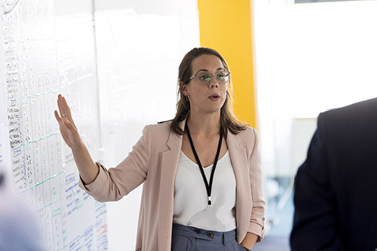 a young woman wearing glasses, explaining something on a white board behind her