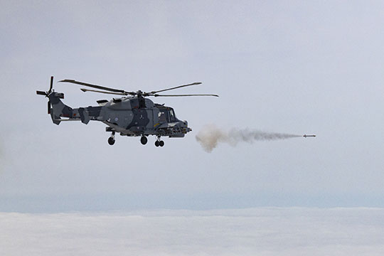 A camouflaged assault helicopter fires a missile which leaves a trail of smoke against a backdrop of grey sky