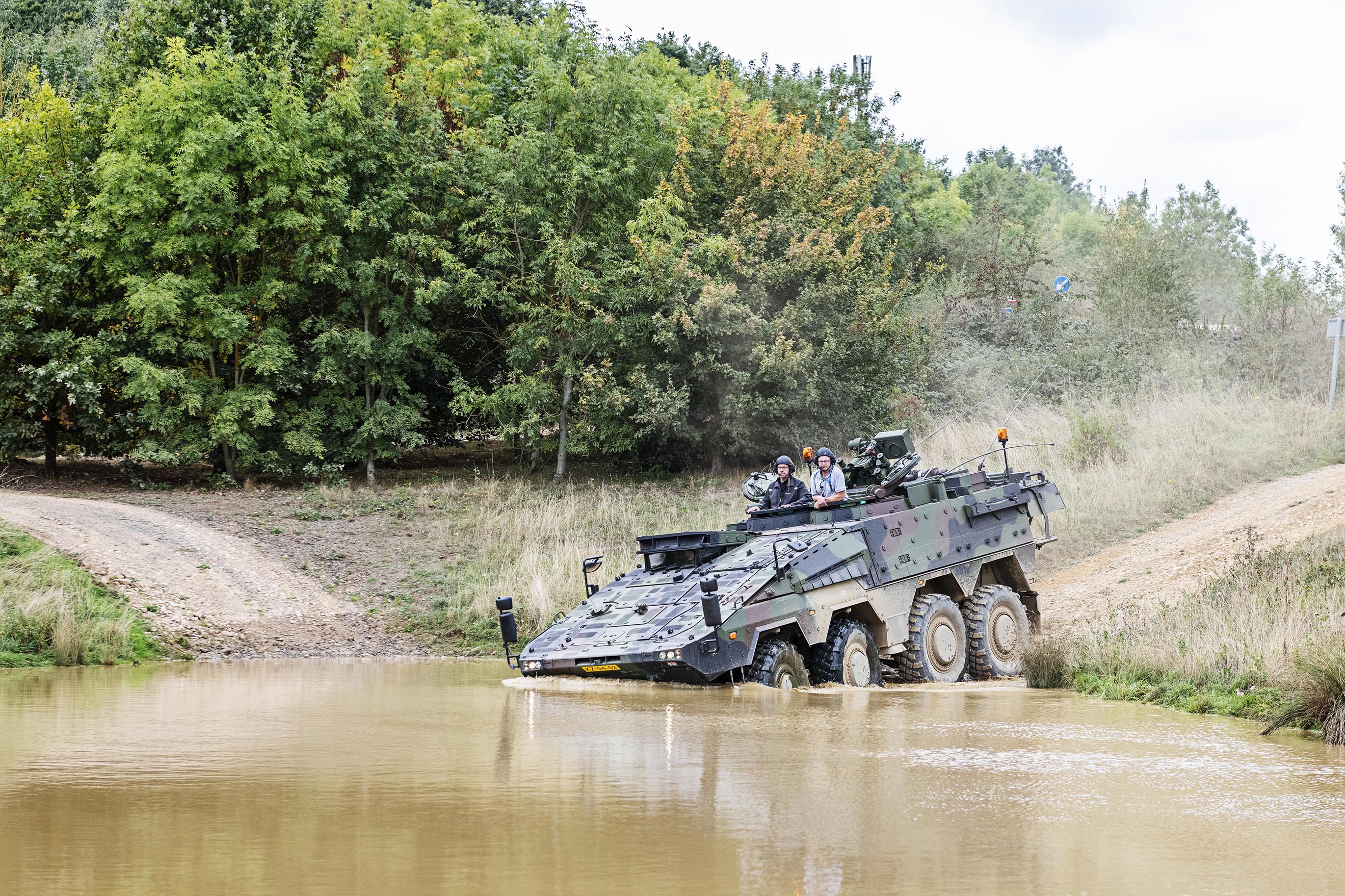 A heavily-armoured, camouflaged tank-like vehicle drives down a hill into a muddy lake, with the front wheels already submerged