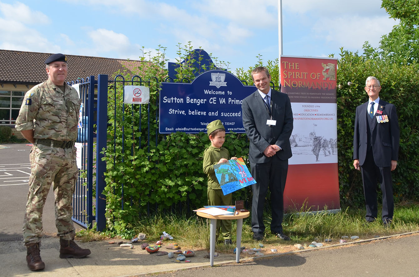 A young child holding a colourful poster celebrating VE Day 75, with two men in suits to his right, and a soldier in uniform to his left.