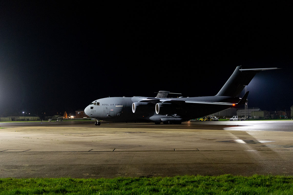 A large grey plane stands on a runway prior to take-off at night, illuminated by floodlights