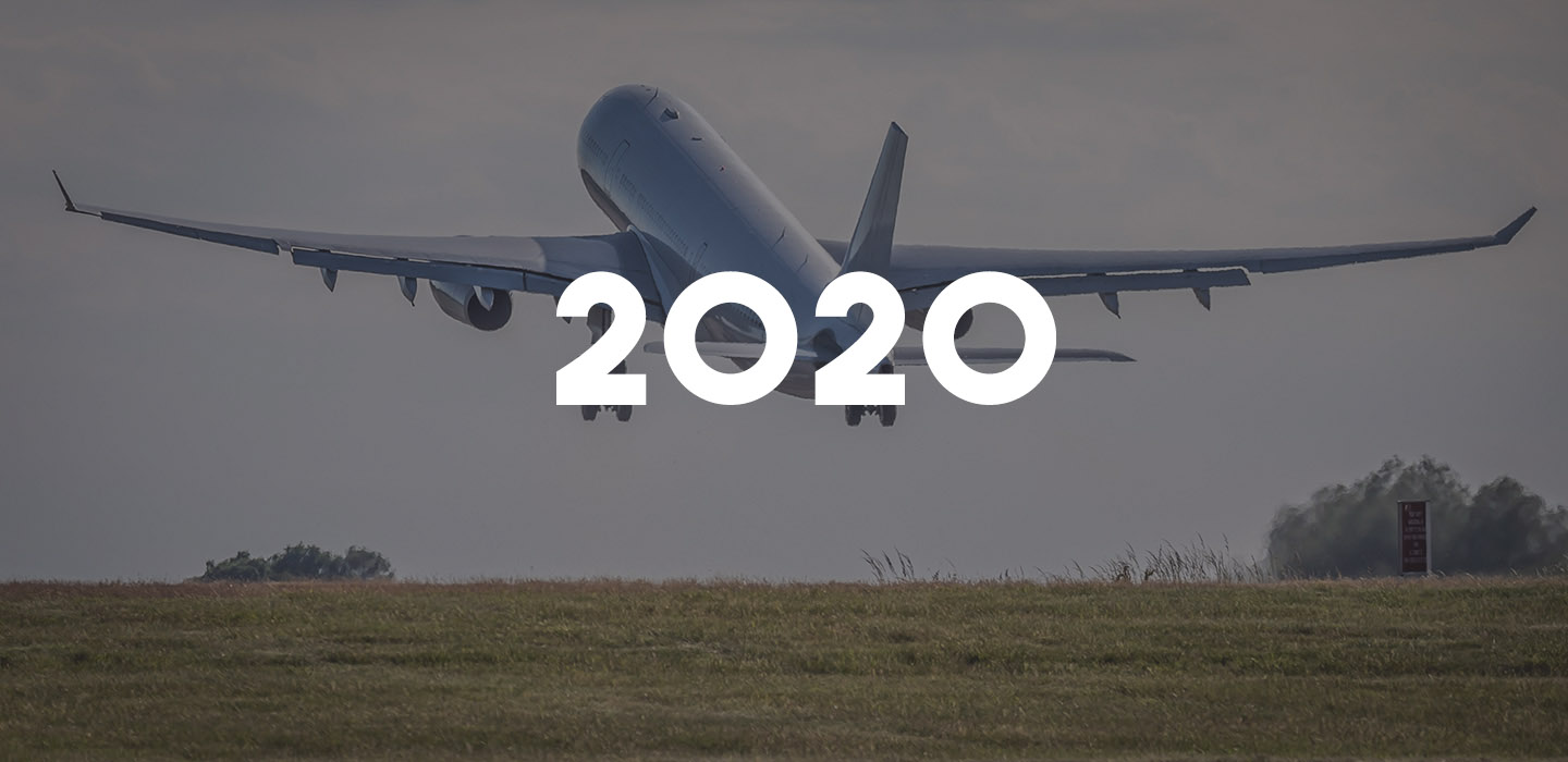 A grey plane climbs into the sky as it takes off from a runway, with "2020" written in the foreground