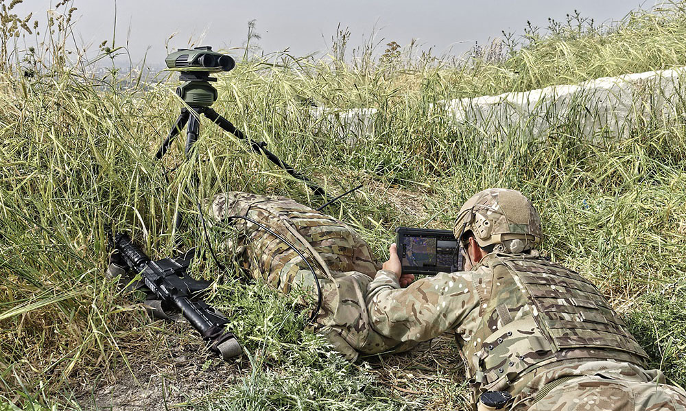 A camouflaged solider lies in the grass, using a laptop and digital binoculars to find their tarrget