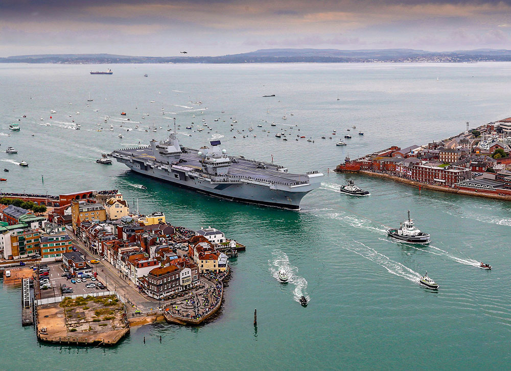 A large aircraft carrier surrounded by small boats comes into harbour with the city in the background