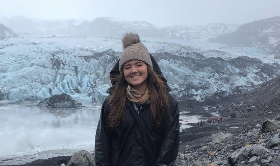 A young woman in a wooly hat and thick winter coat smiles in front of a glacier