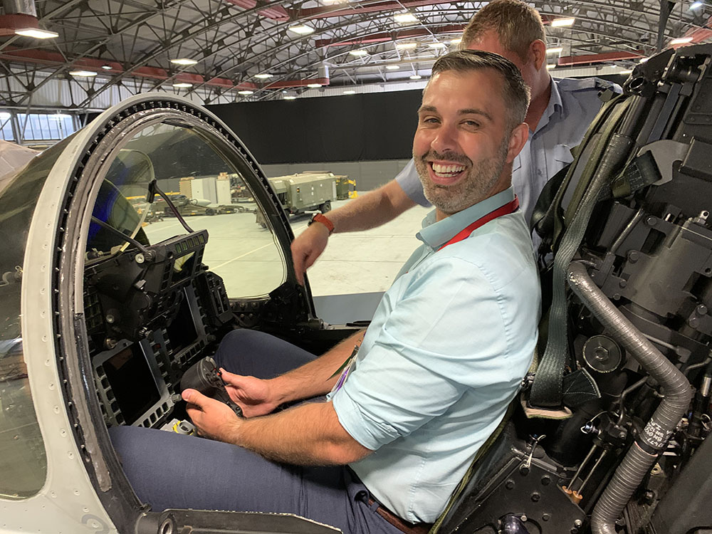A man smiles as he sits in the cockpit of a fighter plane, wearing a light blue t-shirt