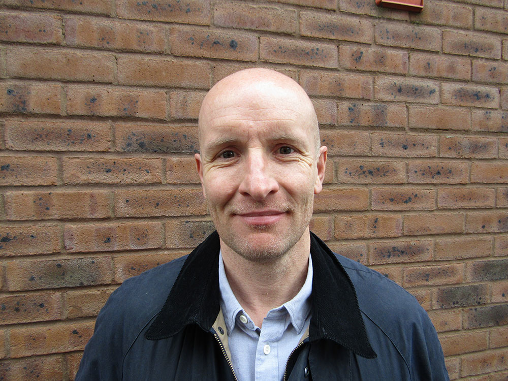 Integrated Logistics Support Manager, Justyn, a bald man in a jacket smiles at the camera with a brick wall behind him