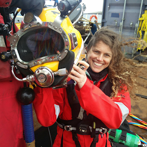 A young brown-haired woman in a bright red diving suit holds a diving helmet aloft as she smiles at the camera