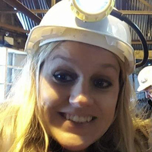 A young blonde woman in a white hard hat with a light on top, smiles at the camera
