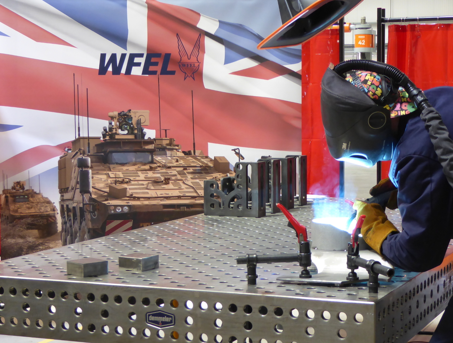 Man in mask produces parts for Boxer vehicle, with Boxer and Union Jack in the background