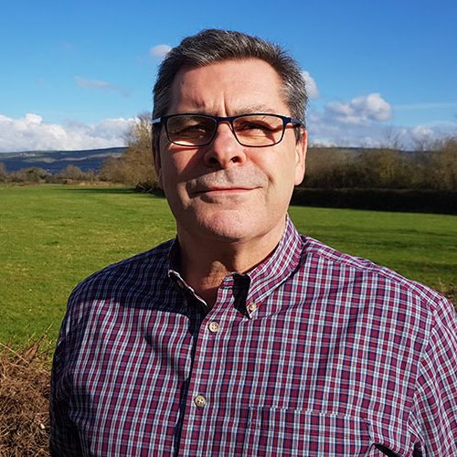 Commercial Manager, Simon, in a check shirt and glasses smiles in the sunshine with a green field and blue sky behind him